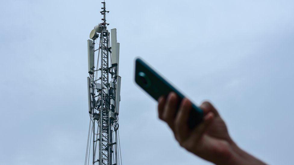 Hand houdt een smartphone omhoog naar een gsm-zendmast tegen een bewolkte lucht.